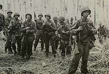 Black and white photo of men wearing military uniforms and carrying rifles standing in front of a dense forest of palm trees