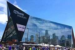 Blue sky, large angular modern building with reflective surface (Minneapolis downtown visible in reflection). Sign on protruding end says "u.s. bank stadium." Crowd of people and vendor tents just visible in foreground