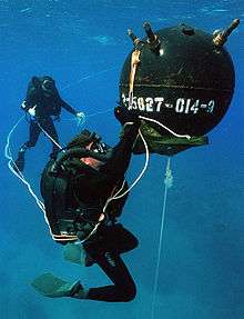  A diver appears to work on a large spherical mine, with another diver observing from a distance in the background