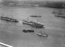 An overhead photograph of eight ships. At the bottom of the photograph is one side of a harbor with the ships in the water. Five ships are displaying decorative bunting. At the top of the photograph is the opposite side of the harbor.