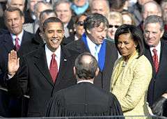 With right hand raised, Barack Obama smiles at a balding man with his back to the camera, as Michelle Obama and others watch