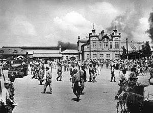A city square filled with soldiers and civilians. Smoke is in the sky in the background behind a large building