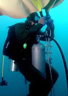 Underwater, near the reef, a US Naval diver in a scuba suit with mask, oxygen tank, and regulator, is attaching a large, upside-down beige bag to braided metal chain.