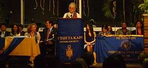 Picture of speaker making speech, with others on podium, with words "Phi Theta Kappa" on banner.