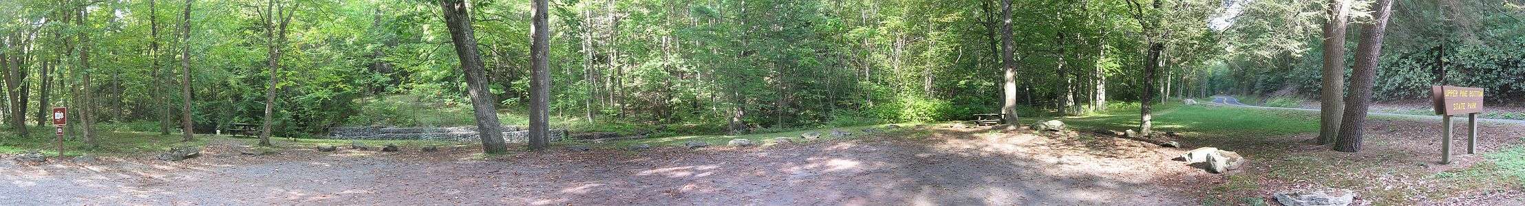 A gravel lot surrounded by grass, large rocks and some large trees is in the foreground, with two picnic tables, the banks of a small stream and dense forest in the background. At the left is a sign reading "Carry in, carry out" and at right is a sign reading "Upper Pine Bottom State Park" with a two-lane highway behind it.