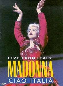 Madonna in short cropped hair, wearing a red flamenco dress. She puts up both of her hands above her head