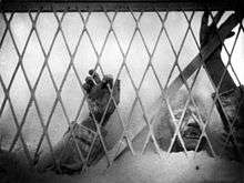 A black and white image of the film depicting the village doctor's hands and arms above flour as he presses up against a chain link fence.