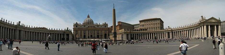 Panorama showing the façade of St. Peter's at the centre with the arms of Bernini's colonnade sweeping out on either side. It is midday and tourists are walking and taking photographs.