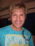 A man in a light blue t-shirt smiles towards the camera.  He has blond tousled hair, and sports a stubble.