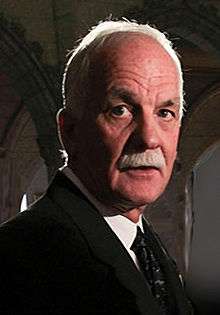 A photograph of a man with white hair and a white moustache facing the right and looking just to the left of the viewer while wearing a necktie