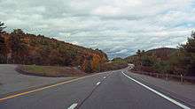 A view of a divided highway from its righthand roadway, with two lanes separated by a dashed white line. There is almost no other traffic; the surrounding area is wooded with some autumn color visible. Ahead the roadways curves by a hill and disappears.