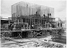 Scaffolding surrounds a half finished concrete foundation. Dozens of metal steel poles rise from the foundation. A dozen workmen are visible and involved in various construction tasks.