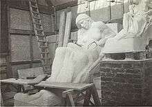 The partially completed statue of a reclined woman sits to the right of a half sized model of the same statue. It appears the work is being conducted inside a temporary structure.