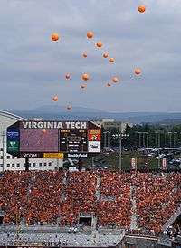 Orange balloons rising above Lane Stadium, with everyone in the stands wearing maroon or orange, and the stadium scoreboard in the background.