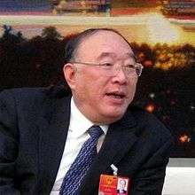 a visibly aged man bending rightwards, wearing glasses, a white shirt, a suit, a dark blue tie and a communist party membership card