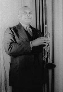 Short-haired African American man wearing a black suit and tie and holding a trumpet, standing facing the camera and smiling.