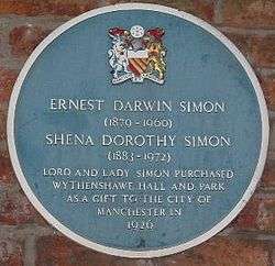 A circular plaque coloured blue mounted on a brick wall, containing the coat of arms of Manchester City Council at the top, the white text "Ernest Darwin Simon (1879–1960)" and "Shena Dorothy Simon (1883–1972)" in the centre, followed by "Lord and Lady Simon purchased Wyhtenshawe Hall and Park as a gift to the City of Manchester in 1926"