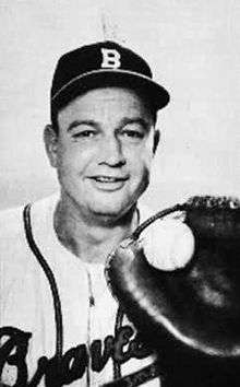 A black-and-white image of a man in a white baseball jersey and dark baseball cap with "B" on the front holding a baseball in a catcher's mitt on his left hand