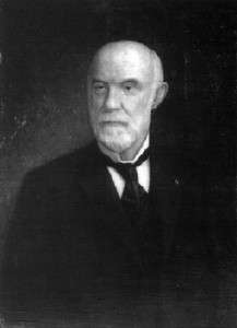 A man in his late fifties, bald on top, with white hair on the sides and a white beard and mustache. He is wearing a black coat and tie and white shirt and facing left