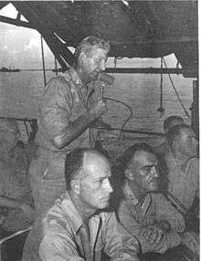 Man standing on deck of ship talking to a microphone. Two other men are seated at a desk in the foreground.