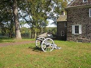 Photo shows an 18th-century cannon, probably a 3-pounder, with an old stone building and the Delaware River in the background.
