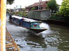 A boat, about ten metres (33 feet) long, travelling along a canal, the dark water breaking up in foam as it passes