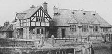 An old photograph showing, from a slight angle, a house on the left and a school on the right.  The house is in stone with a jettied timber-framed upper storey; the school is in stone with three gables in a steep roof containing three dormers and, to the left a bell cote.
