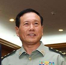 a smiling man with his mouth open and looking rightwards, wearing a light green military uniform with insignia and badges