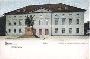 Picture postcard of a large, three-storey building. There is a city square in front of the building. About 10 meters in front of the entrance to the building there is a large bronze statue of two men; the statue is on top of a stone pedestal. The postcard has a white band at its bottom with the printed phrases "Grüss aus Weimar", "Theater", and "499 Verlag von Zedlar und Vogel, Kunstanstalt, Darmstadt 1899."