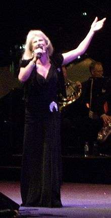 A woman is singing into a microphone held in her right hand, she has her left arm raised with her hand above her head. She wears a dark dress with a jewelled accessory at her left waist. An orchestra in the background is partly obscured.