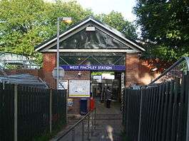 A red-bricked building with a rectangular, dark blue sign reading "WEST FINCHLEY STATION" in white letters all under a light blue sky