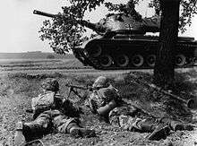 Two soldiers crouch under a tree while a tank sits on a road in front of them.