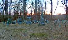 A group of grave markers, some large, some small, seen with bare trees and a dusky sky behind them