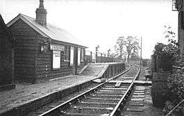 A small wooden hut labelled "Westcott". In front of the hut is a deserted low wooden railway platform with a short section at a much greater height; the only objects on the platform are three large lamps. A single railway track leads past the platform; the line branches immediately past the end of the platform. A cat is asleep on the railway track.
