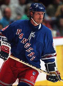 A hockey player stares intently to his left. He is in full uniform with a blue helmet and jersey, and red pants. The jersey has the word "RANGERS" spelled diagonally down his chest.