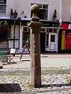 Stone whipping post on paved ground, with shops in the background.