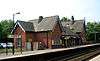 Two sets of rails between platforms beyond which is a brick building with two gables and a canopied area between them; a sign to the left says "Hough Green"