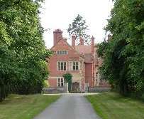 The side of a red-brick house with stone dressings seen along a drive.  On each side of the drive is a lawn and the whole picture is framed by trees.