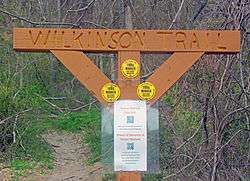 A wooden sign, painted a rusty shade, in the form of a cross with arms holding up the crossbar. On it is carved "Wilkinson Memorial Trail". There is a plastic-covered sign with QR codes below it, and a wooded area to the rear.
