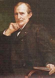 A man with receding brown hair and a mustache wearing a white shirt and black coat and tie. He rests his chin on his right hand and holds a book with his left.