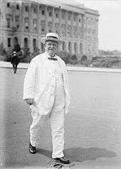 A man in his mid-sixties, wearing a white suit and hat with a black bow tie, walking in front of a building