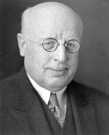 A bald, heavyset white man wearing round-rimmed glasses.