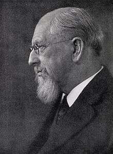 Portrait black and white photograph showing Dall's left profile. Dall's balding head, beard and glasses are shown and is wearing a serious expression on his face. He is wearing a dark coat and suit with a white shirt and a dark tie.