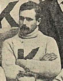 A man with a mustache is looking down to his right and wearing a sweater with a big K on it.