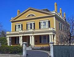 A mustard-yellow two story house with white trim and black shutters has a short semi-circular going to its front. A single story porch with white pillars extends the full width of the house.