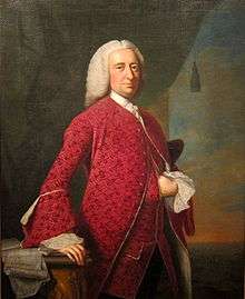 A three quarter length portrait of William Shirley. He wears a red coat over a white shirt with lace ruffled cuffs. His right hand rests on desk or table containing papers.