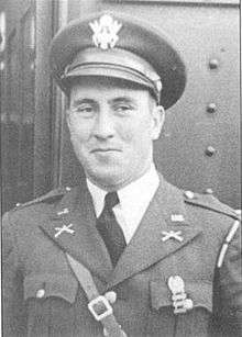 Head and torso of a smirking white man wearing a peaked cap and a military jacket, adorned with pins and badges and a strap running diagonally across his chest, over a shirt and tie.