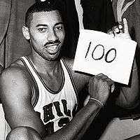 Wilt Chamberlain, an African American man, is shown sitting down in his Philadelphia Warriors jersey while holding up a piece of paper that has the number 100 written on it. The photograph was taken directly after the game and is in black and white.