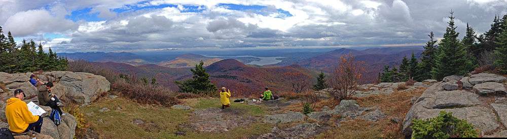 A wide open ledge, with small coniferous trees on either side, on which several people are sitting down and reading, eating or taking photographs. Beyond it lies a mountainous region, with many peaks in fall color. A large body of water is visible just right of center, at the foot of some smaller mountains.