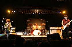 Three men on a stage in front of a crowd; two are holding guitars while the one of the center is sitting behind a drum set. Audio equipment, a drum set, lighting, and other stage fixtures can also be seen in the background.